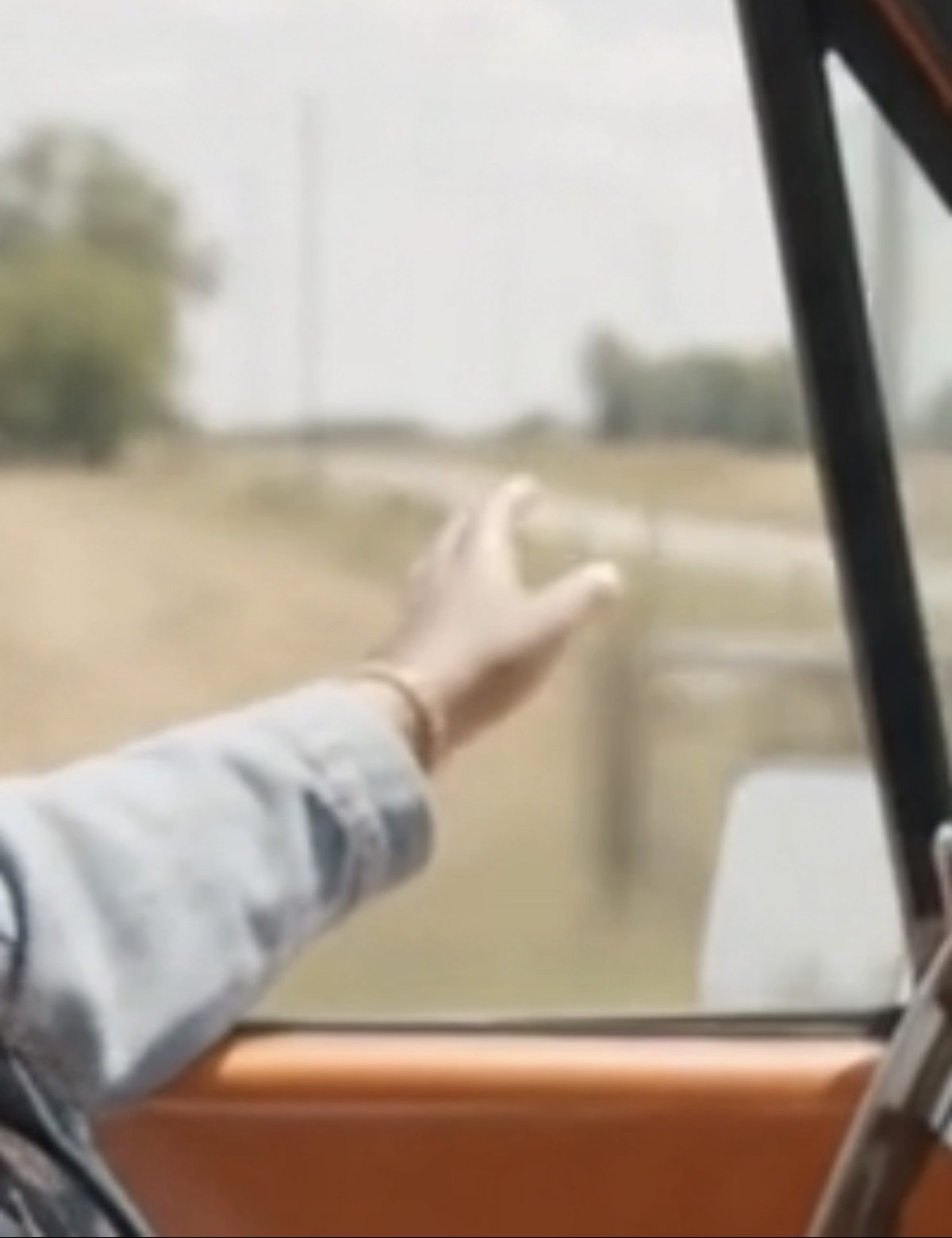 A hand reaching out of a car window over a sun-bleached dirt road, sky wide and pale
